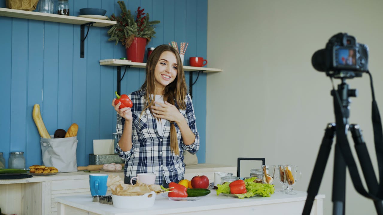 services-03 Young woman in a kitchen holding a bell pepper, preparing food, and recording a video.